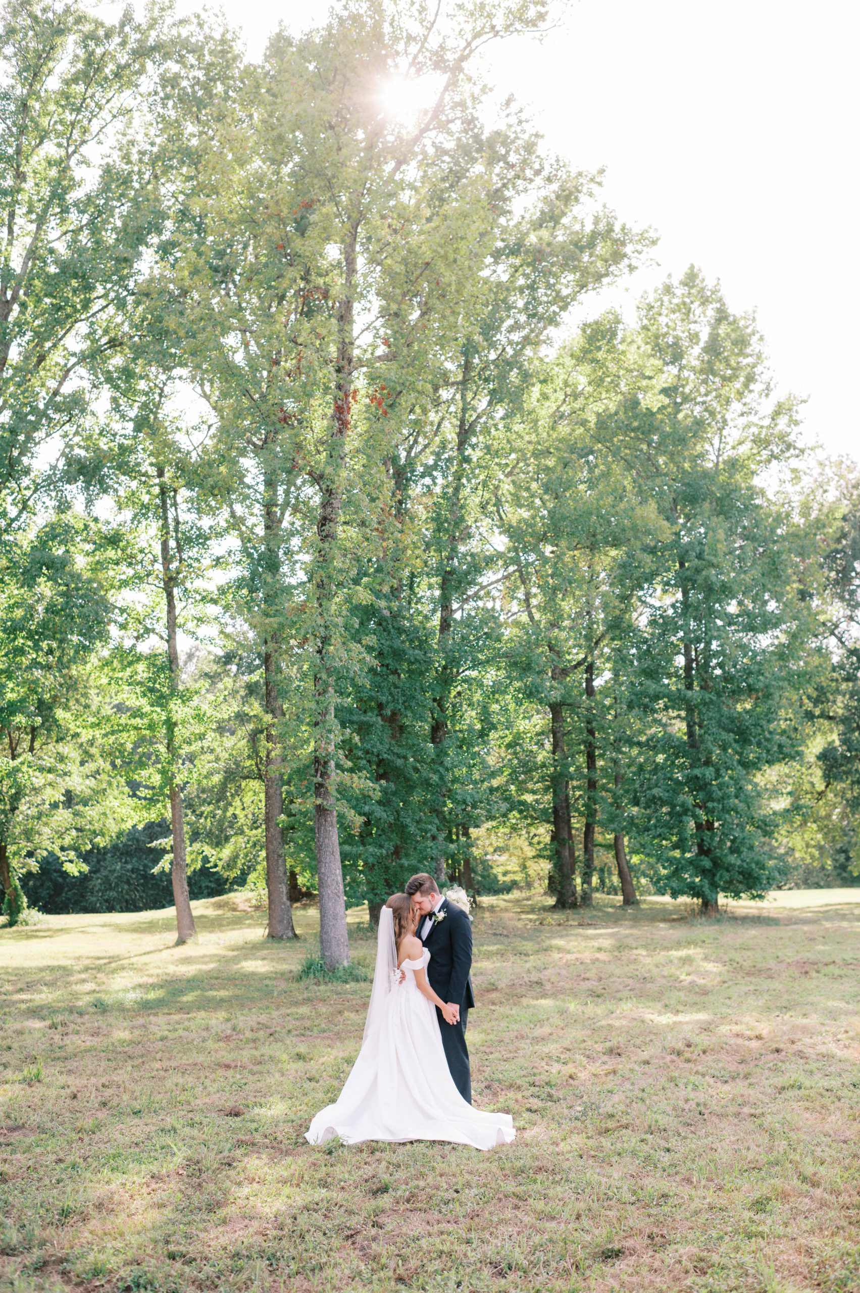bride and groom standing in a grassy forest, facing each other and cuddling together.