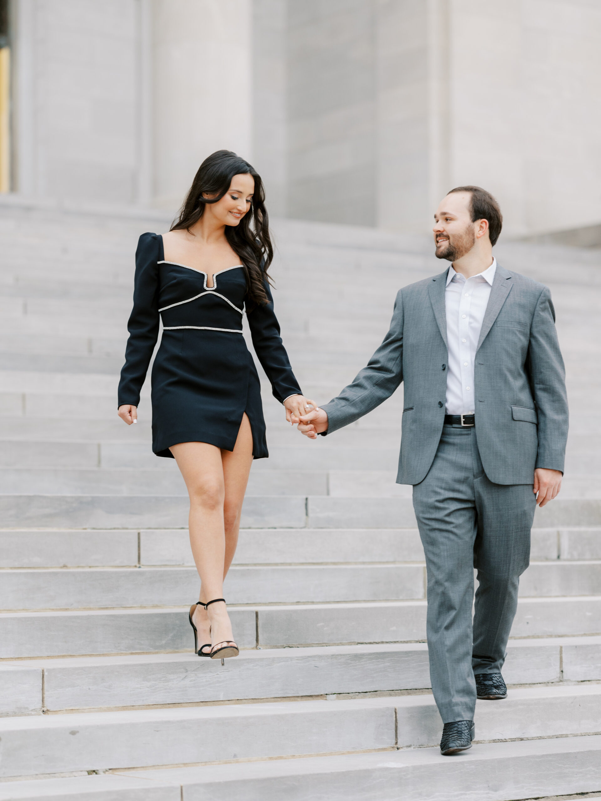 Couple walks down the steps of the Arkansas State Capitol together, wearing a classy, short black dress with black heels and groom wearing a grey suit